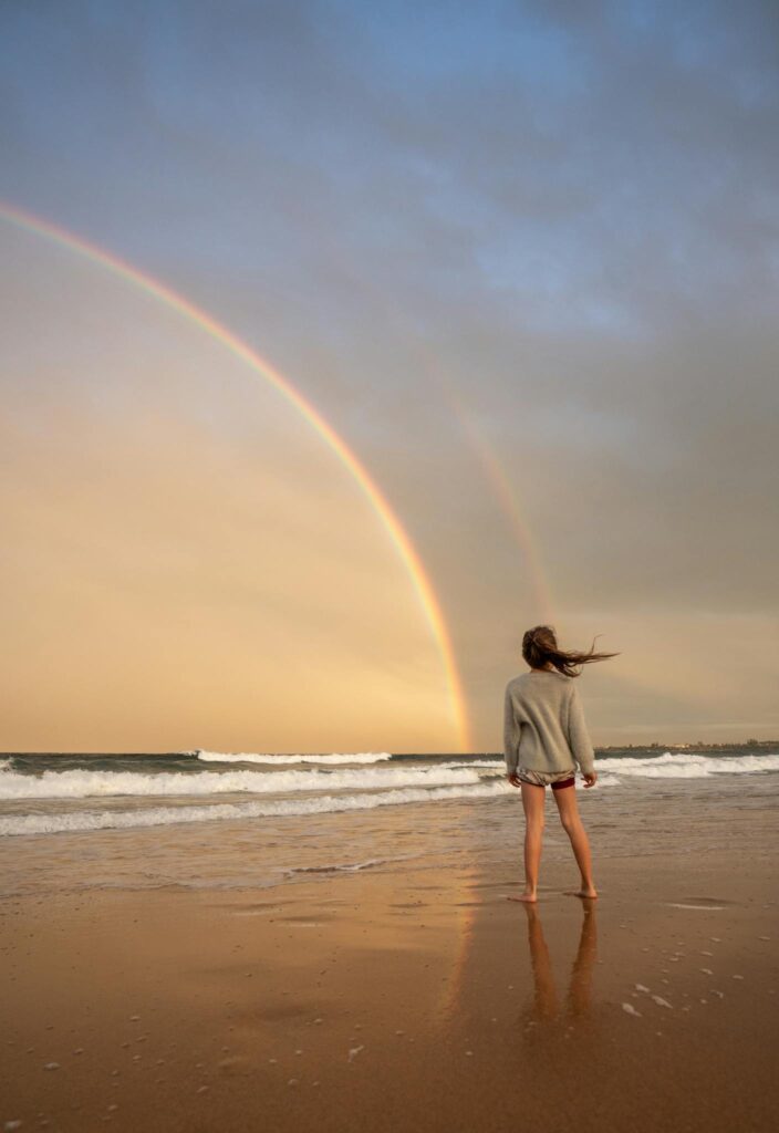 Full body of anonymous barefoot female with flying hair standing on sandy coast near waving sea against colorful rainbow in nature